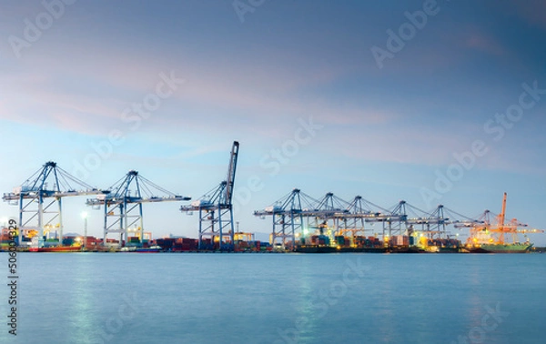 Fototapeta Container cargo ship with crane bridge working in shipyard at dusk.