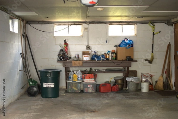 Obraz The interior of an attached car garage on a house, Midwest, USA.  Various  yard keeping tools, cleaning products, a trash can, weed-eater, and miscellaneous other items are seen.
