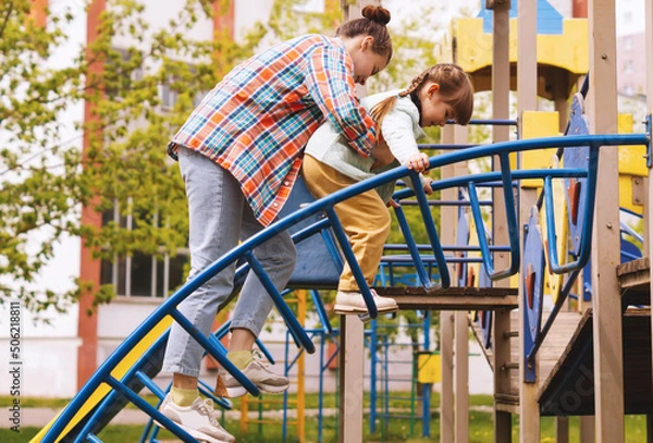 Obraz Sisters play on the playground on a summer day. A white girl in her 20s is playing in the park with a little girl.The older sister helps her younger sister up the stairs by supporting her.Nursing care
