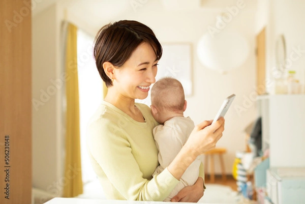 Fototapeta 赤ちゃんを抱っこしながら携帯電話を操作する母親