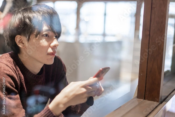 Fototapeta カフェの窓辺でスマートフォンを操作する20代男性