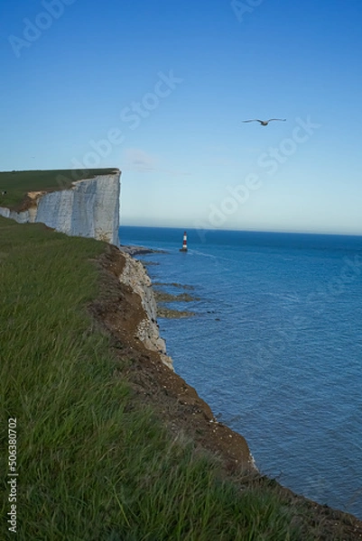 Fototapeta View of the red lighthouse in the bright blue water of the ocean. View of the lighthouse from the top of the cliff. Red lighthouse at the foot of the cliff.