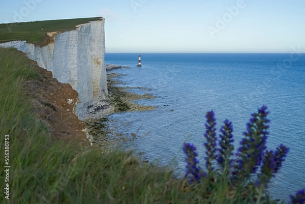 Fototapeta View of the red lighthouse in the bright blue water of the ocean. View of the lighthouse from the top of the cliff. Red lighthouse at the foot of the cliff.