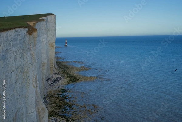Fototapeta View of the red lighthouse in the bright blue water of the ocean. View of the lighthouse from the top of the cliff. Red lighthouse at the foot of the cliff.