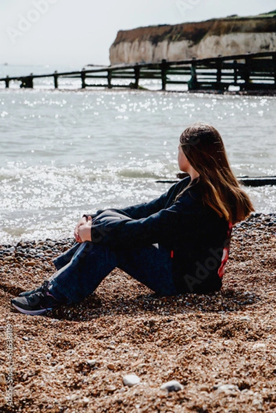 Fototapeta A lonely girl in a black sweater with long hair sits on the beach. A girl sits on the seashore and looks at the sea.