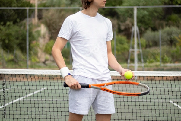 Fototapeta Close-up impersonal portrait of a tennis player holding the ball and a racket by the net on a green tennis court.