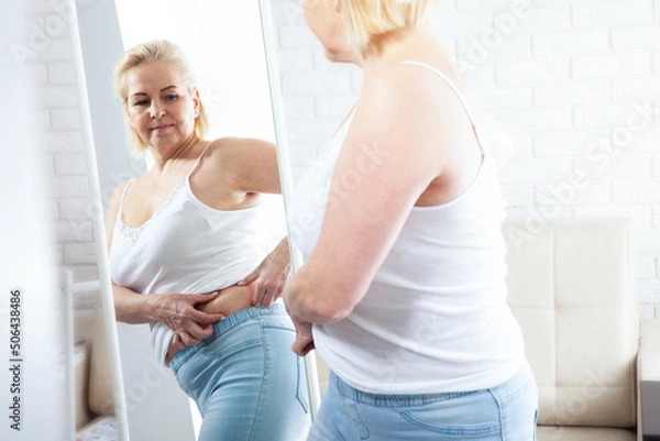 Obraz Displeased middle aged woman looking at herself in the mirror and pinching stomach fat in domestic interior. Sad plus size woman looking her reflection in the mirror with sorrow at home.