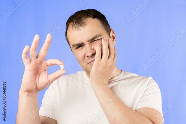 Obraz Man in a white T-shirt on a blue background holds a wisdom tooth in his hands after surgical tooth extraction.Man after an operation to remove wisdom teeth.Pain in wisdom teeth, concept of dentistry
