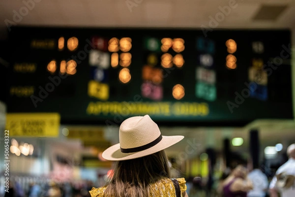 Obraz An attendee at a horse race, wearing a fancy hat in front of horse racing odds.