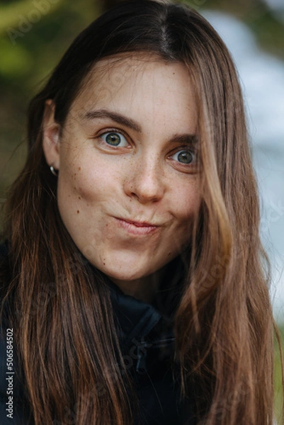 Fototapeta The photo captures a close-up of a young woman making a playful, silly face. Her eyes are wide open, and her lips are pursed, giving her an exaggerated expression. She has long brown hair, and is wear