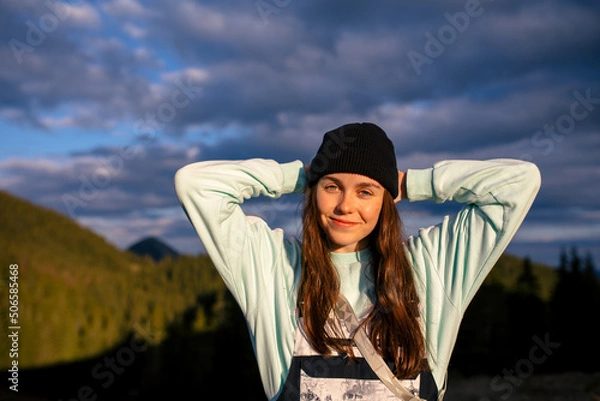 Fototapeta Incredible girl in a black hat stands in the mountains with arms raised against the backdrop of mountains and with a very beautiful sunset