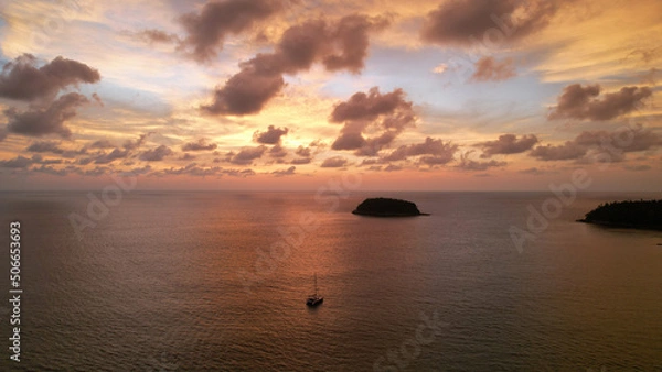 Fototapeta Luxury yacht at an epic sunset with a view of the island and incredible orange clouds. The sky is reflected in the water. A lonely island in the distance. Calm. Top view from drone. Kata Beach Phuket