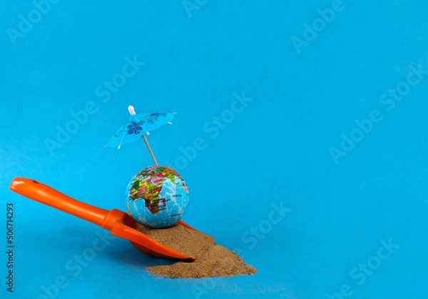 Obraz A globe on the sand under an umbrella from the sun on a blue background. Journeys. Summer holidays