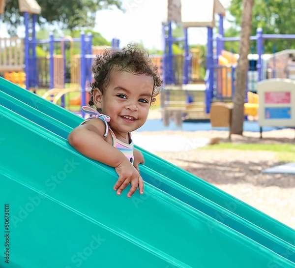 Fototapeta Young girl with curly hair has fun as she goes down a playground slide at a local park. 