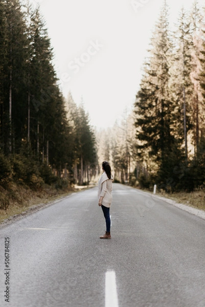 Fototapeta A woman is standing on an empty street in the forest and watching the trees. She is in the middle of the road. Blurry and hazy background, large pine trees. Woodland outdoor traveling. 