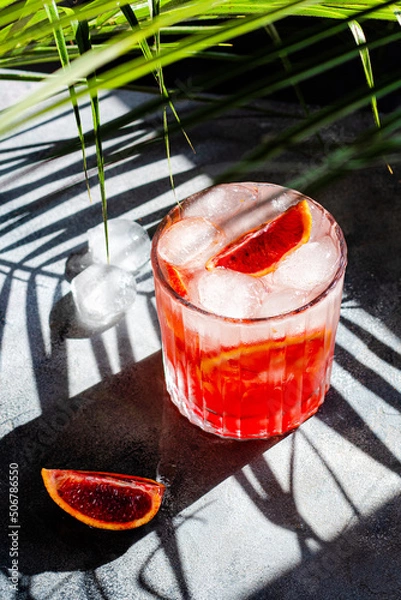 Fototapeta glass with cold mineral water with blood oranges and ice cubes on concrete background. Tropical palm leaf shadow. Sunlight and shadow background.