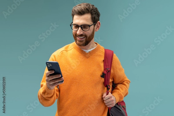 Fototapeta Portrait of cheerful brunette student man with beard using cellphone and smiling, reading good news message, enjoying mobile application. Indoor studio shot isolated on blue background