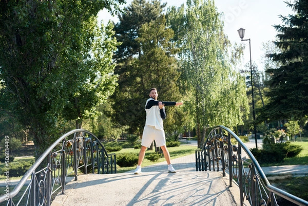 Obraz Young brunette man do morning exercises in city park standing on bridge and wave his hands