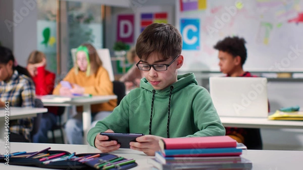 Fototapeta Teenage boy watching video on smartphone sitting at desk in class