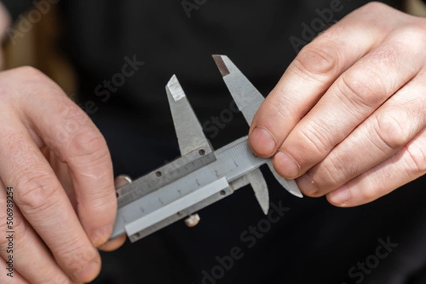 Fototapeta A man's hand holding a caliper. An adult man with a high-precision metal instrument for measuring the external and internal dimensions of objects and the depths