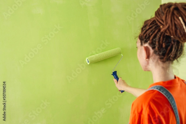 Obraz Woman with dreadlocks, in an orange t-shirt and denim jumpsuit is painting the wall with a roller in green color. Concept of repair, renovation of the new apartment. Copy space