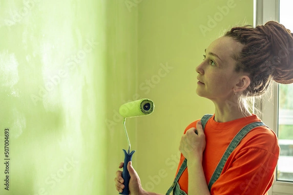 Obraz Woman with dreadlocks, in an orange t-shirt and denim jumpsuit is painting the wall with a roller in green color. Concept of repair, renovation of the new apartment. Copy space
