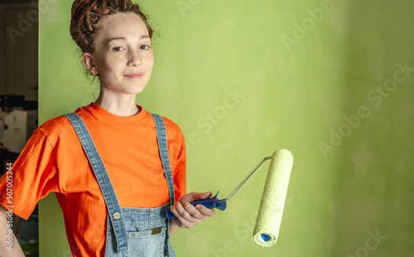 Obraz Woman painter with dreadlocks, in an orange T-shirt and a denim jumpsuit make repairs in the apartment and paints the wall green with a roller