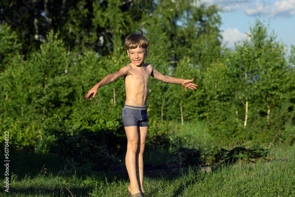 Fototapeta Happy smiling boy in wet shorts plays with water sprinkler on natural blurred background. Summer games on vacation. Happy life.