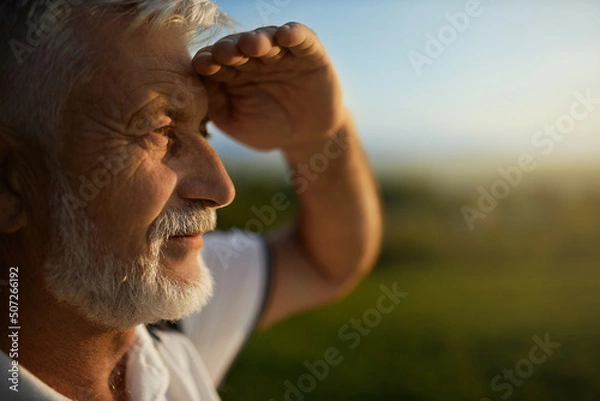 Fototapeta Senior bearded man using hand to protect eyes from sun, smiling outside. Close up view of male tourist squinting from bright light, while looking into distance, during mountain trip. Concept of watch.