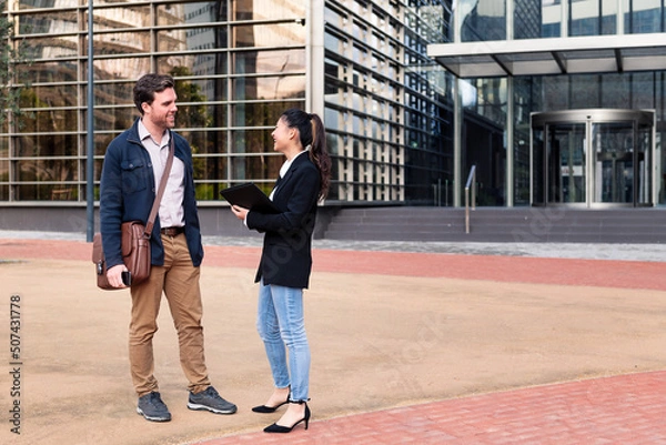 Fototapeta caucasian man and asiatic woman smiling and talking about business in front of an office building, concept of coworkers and lifestyle, copy space for text