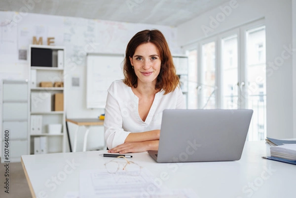 Obraz young woman sits in the office in front of her laptop and looks into the camera