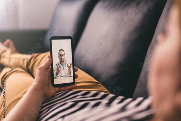 Obraz Woman lying on a sofa and talking with a doctor online using mobile phone. Telemedicine concept.