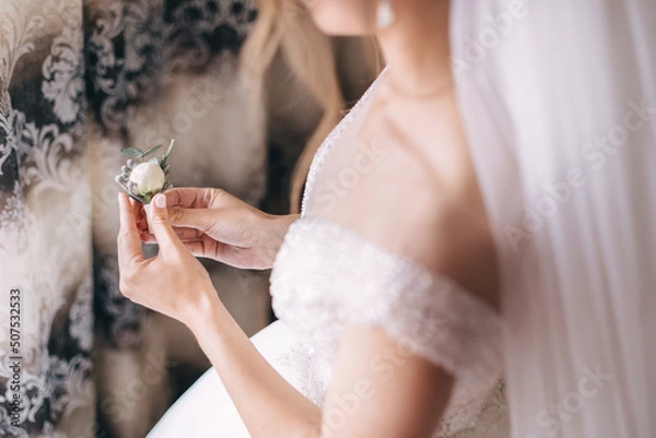 Fototapeta The bride holds a boutonniere with a white rose in her hands, waiting for the groom.