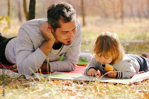 Fototapeta Father and son reading book lying on green grass in park. Concept of happy family relations and carefree leisure time