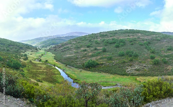 Fototapeta Panoramic of a river running through the meadow in a green valley with mountains in the background.