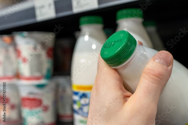 Fototapeta Buyer checks expiration date of dairy product before buying it. Hand holding milk bottle in supermarket.