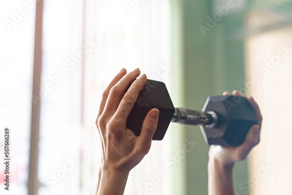 Obraz Side view of fit adult slim woman lifting weight in fitness box. Close-up on the arm with muscles in front of the window. Workout at home or intdoors. Only hands