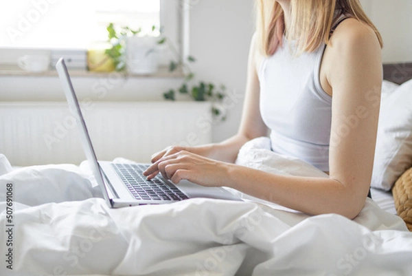 Fototapeta Happy casual beautiful woman working on a laptop sitting on the bed at the morning in the house. Working at home, freelance concept.