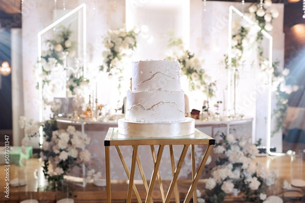 Obraz The wedding cake. White, two-tier, decorated with gold. On a delicate light background and a golden stand. Side view.
