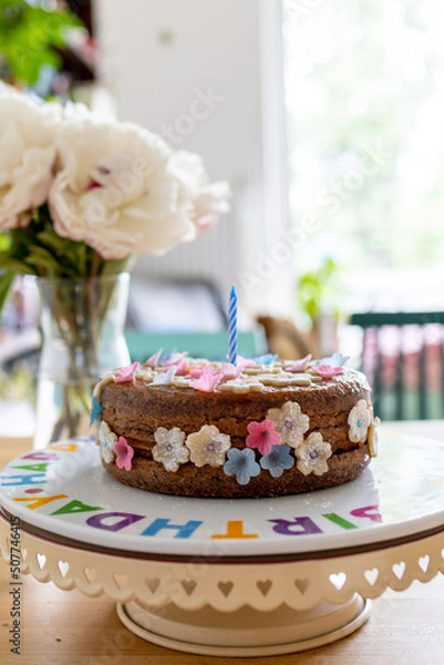 Fototapeta Birthday cake decorated with marzipan flowers on a decorative birthday plate with flowers and a window in the background