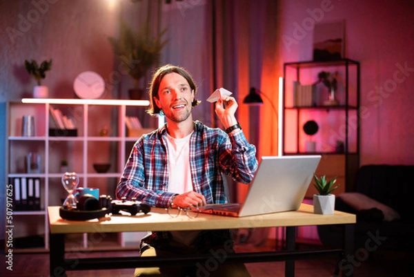 Fototapeta Happy young man in casual wear throwing paper plane while taking break during remote work on laptop. Smiling caucasian male sitting at desk during evening time and having fun.