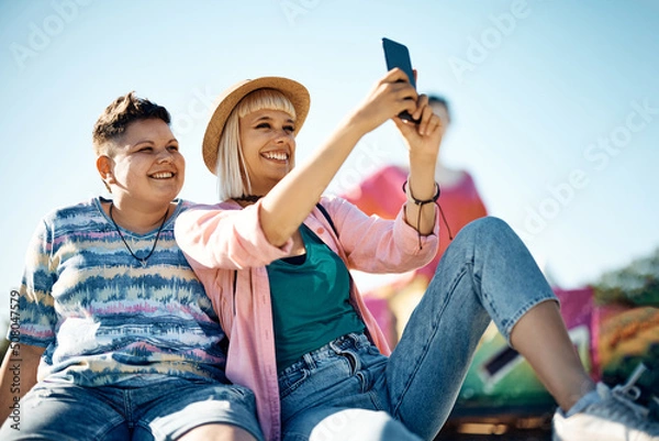 Fototapeta Happy female friends taking selfie while having fun at amusement park.