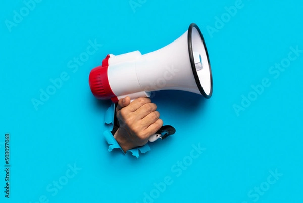 Fototapeta hand holds a megaphone blue background out of a torn hole in a paper background