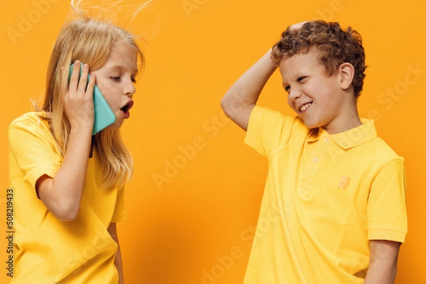 Fototapeta cute, cheerful children, brother and sister, of school age in yellow clothes stand on an orange background and the girl is enthusiastically talking on the phone.
