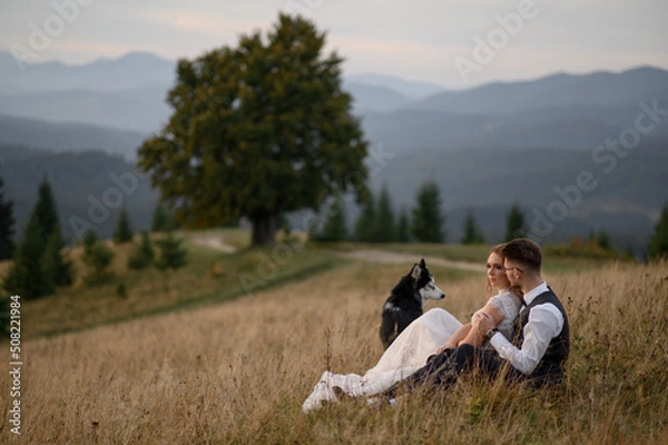 Fototapeta wedding couple with dog in mountains at sunset. romantic evening in mountains.