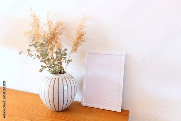 Fototapeta Blank photo frame, eucalyptus and dry reed in a vase. Modern Scandinavian style interior on the beautiful chest of drawers with white wall and bright room. Elegant interior. Summer, fall, still life.