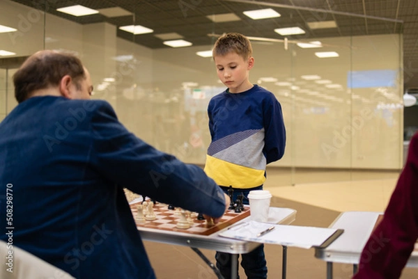 Fototapeta Concentrated boy develops a chess strategy, plays a board game with adult professor .Chess game in action. Board in details. 