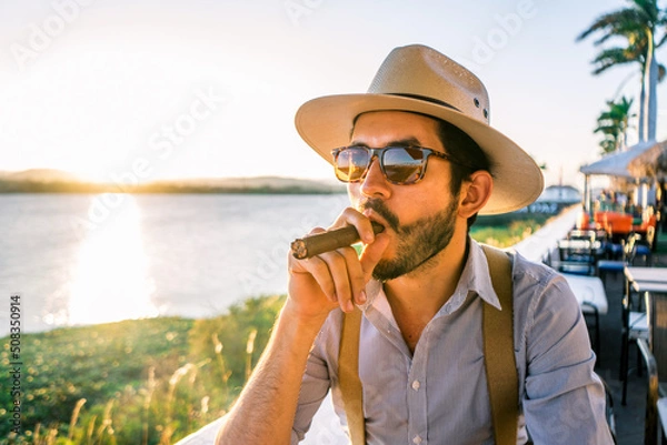 Fototapeta Latin hipster in a bar on the shore of a lake at sunset wearing a hat and suspenders and smoking a cigar in Managua Nicaragua