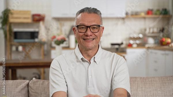 Obraz Portrait of happy senior asian man smiling at home. Old japanese man relaxing on sofa and looking at camera. Portrait of elderly man enjoying retirement.