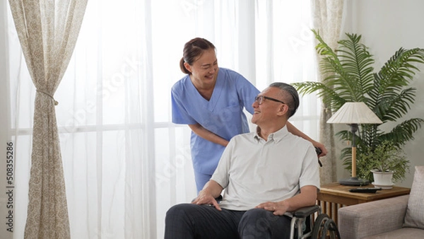 Fototapeta Friendly taiwanese woman nursing aide putting hands on disabled grandfather’s shoulders while talking to him at home. he sits on a wheelchair and has smile on face